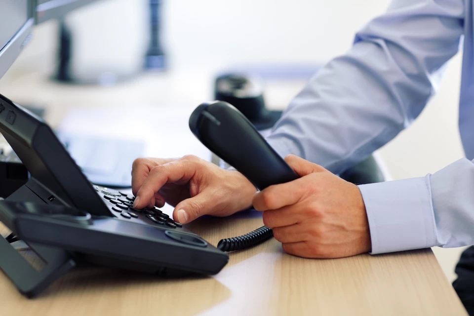 Un professionnel en chemise claire compose un num&eacute;ro sur un t&eacute;l&eacute;phone fixe de bureau dans un environnement de travail moderne
