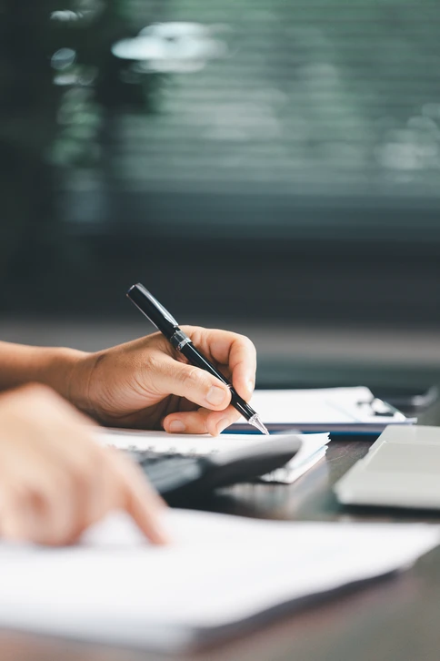 Photographie d'un professionnel en costume assis à son bureau dans un cabinet comptable moderne, étudiant attentivement des documents financiers (bilans, rapports comptables) éclairés par la lumière naturelle d'une fenêtre. Sur le bureau sont visibles un ordinateur, des classeurs et des documents professionnels. L'expression concentrée du professionnel suggère l'analyse minutieuse et la rigueur dans la gestion comptable. Arrière-plan professionnel avec bibliothèque ou mobilier de bureau discret.